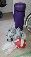 Photo showing two purple yoga mats rolled and secured with black straps, pair of blue and white Celena ice skates size 11 with black laces, Molten red and white volleyball, white frisbee, and two white Asics knee pads on carpet.