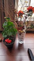 Photo showing Mary Kay crystal vase filled with faux orange-red flowers, decorative dried branches, and small colored eucalyptus leaves, alongside a black plastic pot with live potted geranium plant with red flowers.