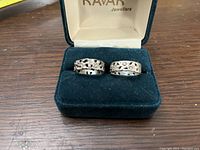 Two antique sterling silver cigar band style rings with pierced floral and vine designs displayed in a black velvet jewelry box under the lid marked Kavar Jewellers.
