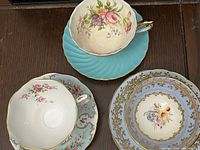 Photo showing three bone china teacups and saucers stacked on a wooden surface, illustrating floral patterns and pale blue colors with gold accents.