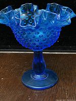 Front view of cobalt blue hobnail textured pedestal vase with ruffled edges, placed on dark surface against dark background