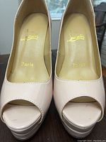 Close-up top view of left and right nude patent leather pumps showing gold script marks inside reading 'Louboutin Paris', peep toe front and slight wear marks on toes.