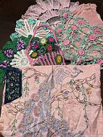 View of assorted crocheted doilies and hand-embroidered textiles in pastel colors from 1950s and 60s, showing floral and lace patterns and a peacock embroidery.