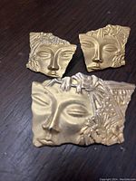Photo showing three pieces: one larger pin and two matching earrings designed as stylized faces in gold-tone metal with floral details.