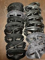 Top view of 20 black masquerade half masks arranged in two columns on a wooden surface, showing cloth-like and plastic finishes with elastic straps