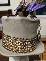 Grey felt top hat with copper-colored beaded and sequined band, purple and gold dragon figure on top, and pocket watch chain visible.