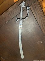Full length view of the curved metal performance sword lying on dark wood surface showing jewel-studded embossed blade and dragon head handle.