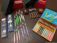 Overview photo showing three sets of retro mini forks with colorful resin handles in original packaging, two collector souvenir spoons in cases, and wooden salt and pepper shakers on table.
