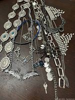 Various silvertone costume jewelry pieces spread out on a wooden surface, featuring bat necklaces, faux pearl drapes, coin belt, shield belt with turquoise stones, and link necklace.