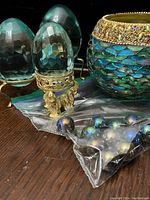 Photo shows three varying sized turquoise glass eggs including two faceted and one smooth, each on ornate brass stands, the iridescent glass candle holder with turquoise and purple hues, and a bag of iridescent purple/black marbles.