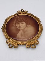 Photo showing the front view of the gilded metal frame with a sepia-tone portrait of a woman inside. Ornamentation includes floral decorations and scrollwork at top and bottom.