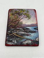 Front view of the miniature landscape painting showing rocky shoreline, water, trees, and sky with signature visible in paint.