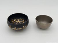 Two singing bowls side by side, one decorated dark bowl with gold script and one plain metal bowl
