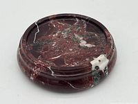 Round solid stone base with polished dark reddish-brown surface with white and green veining, circular groove on top, small white patch on side.