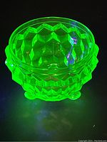 Uranium glass dish fluorescing bright green under UV light, showing geometric diamond-cut pattern and three small feet