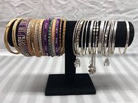 Full view of 23 bracelets/bangles displayed on a black velvet stand, showing gold-tone, silver-tone, and beaded bangles with decorative details.