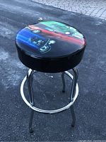 Side angled view of the garage stool showing the motorcycle graphic on the black padded seat and the chrome metal legs with footrest ring.