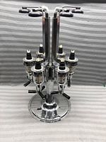 Front view of the chrome metal liqueur dispenser showing four pourer valves with black plastic lever handles and white/black pour spouts on each valve.