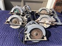 Group photo showing five electric circular saws laid out on a padded blue cloth, three with Skilsaw branding and one Craftsman, all showing rust and dust.