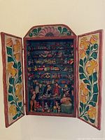 Front view of open wooden wall-mounted box with floral painted doors revealing interior shelves filled with miniature carved and painted toys and figures.