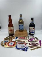 Full lot photo showing two empty glass beer bottles, a solid brass Molson Stock Ale bottle on a wooden pedestal, and a selection of colorful unused Molson beer labels spread in front.