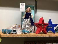 Wide view of two Winter Velvet Barbie dolls, five blue Champion metal cars, Barbie watch in red star-shaped case, Barbie Statue of Liberty blue star-shaped box, and eight composite mini Barbie ornaments on shelf.
