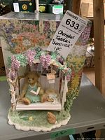 Photo showing Cherished Teddies 'Genovieve' figurine with pergola and swing, detailed floral arch, bear in green dress, small bunny, and straw hat. Box with floral artwork in background.