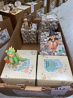 Box containing multiple Cherished Teddies figurines in original packaging showing variety of characters and costumes including dinosaur and wizard outfits.
