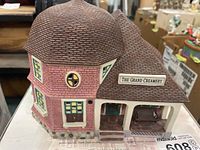 Side and front angled view of The Grand Creamery miniature building showing brown tiled roof, pink bricks, stone foundation, green windows and porch area with bench.