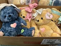 Four plush bears in a cardboard box showing blue, cream, yellow, and light yellow bears with various colored ribbons and pink heart patches on chests