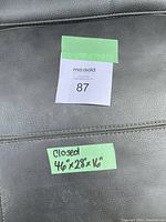 Close-up photo of black vinyl upholstery with stitched detail and two green tape labels showing MaxSold tag and closed dimension label 46"x28"x16".