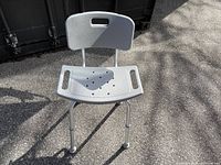 Front and side view of grey plastic height adjustable shower seat with ventilated seat surface and backrest, standing outdoors on asphalt.