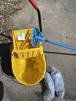 Yellow industrial mop bucket with attached blue wringer, one mop handle, and two new mop heads placed beside it on pavement.