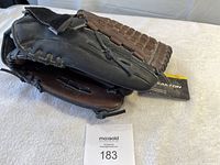 Photo of the black and brown leather baseball glove showing the front side with braided laces and tags attached, placed on white cloth background.