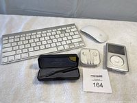 Top view showing the white Bluetooth keyboard missing one key and the battery compartment lid, white Apple Bluetooth mouse, Apple iPod with visible screen, Apple earphones in plastic case, and Lee Valley Space Pen with case
