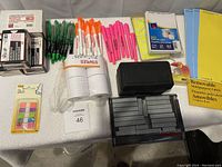 Photo showing assorted pens, unopened Post-it flags, black office supplies like tape dispenser and storage box, several packs of laminating pouch boxes, and removable labels.