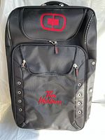 Front view of black cloth suitcase showing red embroidered Tim Hortons logo and stylized upper logo, featuring two front zippered compartments and metal grommets.