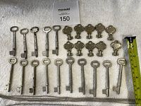Full layout of 15 skeleton keys of two different types plus 10 smaller keys and metal ball chains on a white fabric background with a measuring tape showing length.