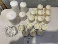 View of 13 pillar candles in white and cream colors arranged on a white fabric surface with a clear glass votive candle holder at front left.
