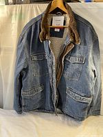 Front view of Wrangler blue denim sherpa-lined jean jacket with brown corduroy collar, chest pockets, and lower pockets with flap closures, displayed on hanger.