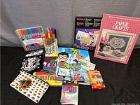 Wide view of all art supplies including books, coloring sheets, crayon boxes, markers, and pencil case on black surface