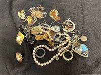 Assorted men's and women's jewelry pieces laid out on a dark fabric background showing various earrings, rings, and necklaces including pearl style beads.