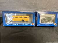 Two boxed Bachmann HO scale motive power vehicles: yellow school bus and green 1950s 4x4 SUV, both with high trailers.