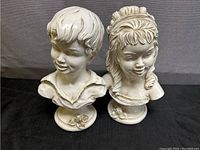 Full view of both plaster busts of a boy and girl on round bases with flowers.