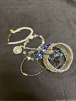 Top view of assorted costume bracelets on dark fabric surface showing various styles and colors including white beads, metallic cuff, and blue accents.