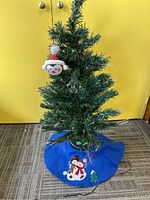 Full view of small artificial fiber optic Christmas tree with blue tree skirt and penguin ornament hung on the tree.