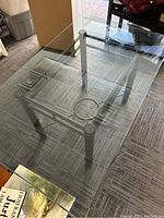 Top view showing clear glass tabletop with slight reflections, four cylindrical iron legs, and metal circular braces below the glass.