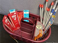 Overall view of burgundy yarn basket containing knitting needles in packaging, orange scissors, and measuring tape