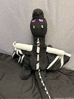 Front view of the Minecraft Ender Dragon plush showing its black fabric, pixelated purple eyes, wings and tail with white spike details.