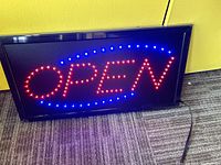 Photo showing the OPEN sign lit with red and blue LEDs, placed on carpet against a yellow wall. The sign is displaying the word OPEN with surrounding blue lights.